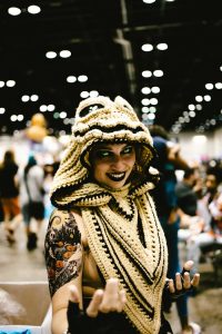 Woman in knitted headwear with tattoos smiles at a bustling comic convention